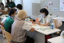 明るい室内で、マスクとエプロンを着用した専門家が歯の模型を手に持ちながら、テーブルを挟んで座る高齢女性たちに熱心にブラッシング指導や口内ケアの説明を行っている、歯科保健指導の様子を捉えた写真