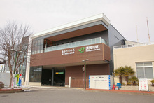 薄いグレーと茶色の外壁を組み合わせたモダンなデザインのJRの駅舎が、曇り空の下に佇んでおり、建物には駅名とともに他の文字も掲げられている、落ち着いた雰囲気の駅前風景を捉えた写真