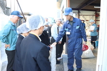 衛生キャップとマスクを着用した学生たちが、青色の作業服を着た指導員の説明を真剣な表情で聞きながら、積み上げられた白い製品を間近で見学している、工場内での校外学習の様子を捉えた写真