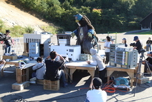 屋外の撮影現場にて、精巧に作られたミニチュアの建物セットが並ぶ中、着ぐるみ姿の怪獣キャラクターを囲んでスタッフたちが機材の調整や演出の確認を行っている、特撮映画のメイキング風景を捉えた写真