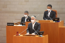 議場の演壇でスーツ姿の人物がマスクを着けて発言しており、背後には木製の机に座る議会関係者と書類が並ぶ市議会定例会本会議の写真