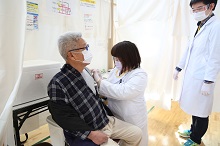チェック柄のシャツを着た高齢男性が、別のスタッフが傍で見守っている中、白い診察衣とマスクを着用した医療スタッフから左腕にワクチンの接種を受けている様子を捉えた写真