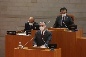 議場の演壇でスーツ姿の人物がマスクを着けて発言しており、背後には木製の机に座る議会関係者と今期定例会で補正予算議案について討論している写真