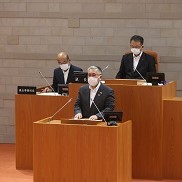 議場の演壇でスーツ姿の人物がマスクを着けて発言しており、背後には木製の机に座る議会関係者と書類が並ぶ市議会臨時会市長挨拶の写真