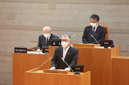 議場の演壇でスーツ姿の人物がマスクを着けて発言しており、背後には木製の机に座る議会関係者と書類が並ぶ市議会臨時会市長挨拶の写真