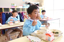 水色のスモックを着た男の子が、教室で箸を使い給食のおかずを口に運びながら、カメラに向かって満面の笑みを浮かべており、背景には同じように給食を食べる他の園児たちの姿が映っている写真