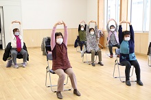 明るい屋内のフローリングの部屋で、マスクを着用した数名の高齢男女が椅子に座り、両手を頭の上で組んで高く伸ばしながらストレッチ体操に励んでいる様子を、斜め前方から捉えた写真