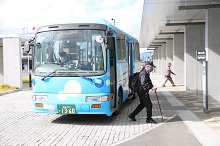 水玉模様が描かれた明るい青色の小型バスが停留所に停車しており、リュックを背負い杖をついた高齢男性が、マスクを着用してゆっくりとバスから降車している写真