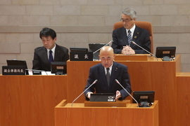 演壇に立ち答弁を行うスーツ姿の男性と、その後方の席に座る議長などと思われる二人の男性が議場に並び、厳かな雰囲気の中で会議が進行している様子を捉えた写真