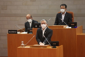 議場の演壇でスーツ姿の人物がマスクを着けて発言しており、背後には木製の机に座る議会関係者と市議会定例会本会議の写真