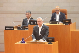 演壇に立ち答弁を行うスーツ姿の男性と、その後方の席に座る議長などと思われる二人の男性が議場に並び、厳かな雰囲気の中で会議が進行している様子を捉えた写真