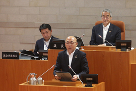 演壇に立ち答弁を行うスーツ姿の男性と、その後方の席に座る議長などと思われる二人の男性が議場に並び、厳かな雰囲気の中で会議が進行している様子を捉えた写真