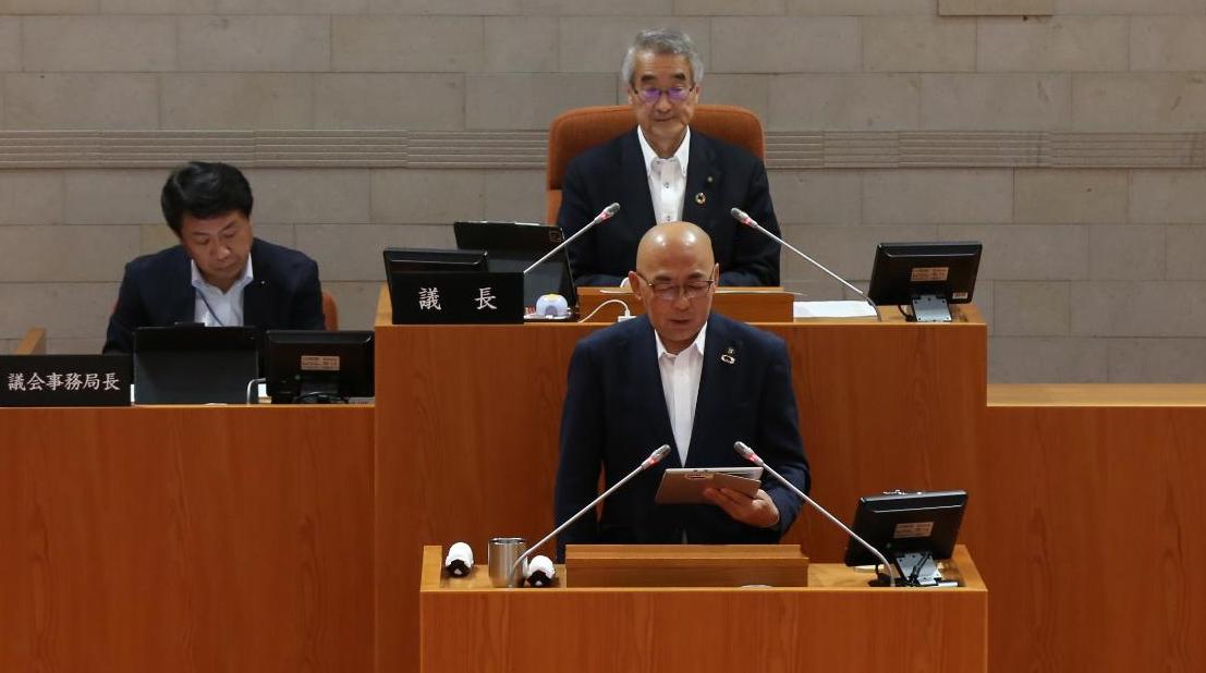 演壇に立ち答弁を行うスーツ姿の男性と、その後方の席に座る議長などと思われる二人の男性が議場に並び、厳かな雰囲気の中で会議が進行している様子を捉えた写真
