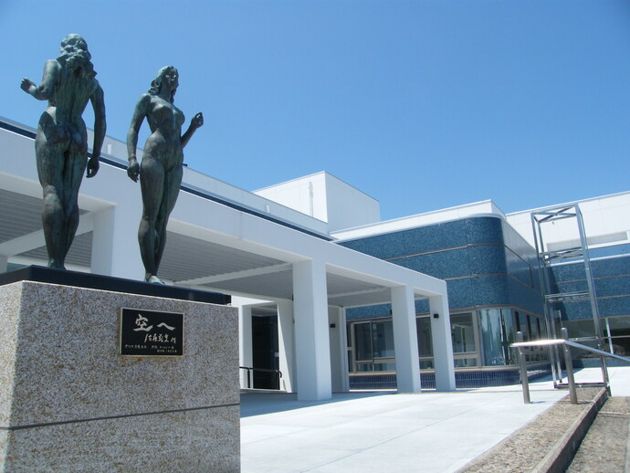 雲一つない青空の下、白を基調とした近代的な文化センターの入り口付近に、佐藤忠良作「空へ」と記された銘板付きの台座に立つ二体の女性のブロンズ像が、建物の開放的なエントランスを見守るように設置された風景を捉えた写真