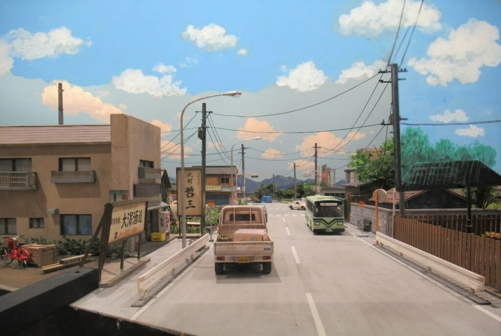 入道雲が浮かぶ青空の下、商店が並ぶ道路を走る軽トラックや路線バス、電柱や街灯が細部まで作り込まれているミニチュアセットの写真