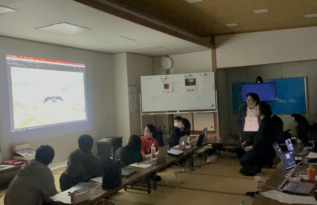 薄暗い和室のワークショップ会場で、壁に投影された飛行怪獣の映像を学生たちが真剣な眼差しで見守り、講師がプロジェクターの横で解説や講評を行っている、特撮映像作品の試写会や最終チェックの様子を捉えた写