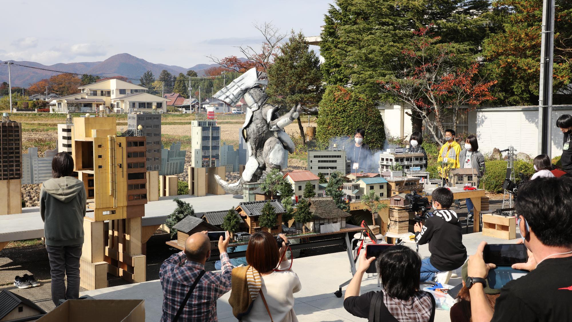 屋外に並ぶ精巧な都市模型の中央に巨大な怪獣像が立ち上がり、その周囲で多くの来場者が撮影や観察をしている様子を捉えた写真