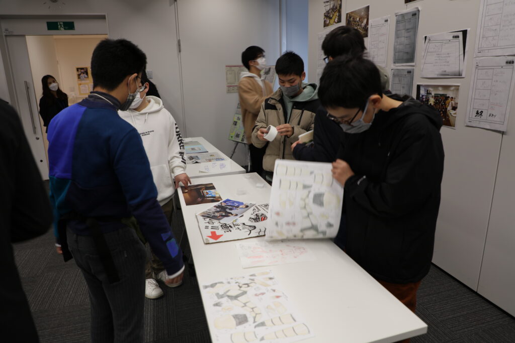 室内で複数の若者たちが白い長机を囲み、手元の資料やイラストを見ている写真