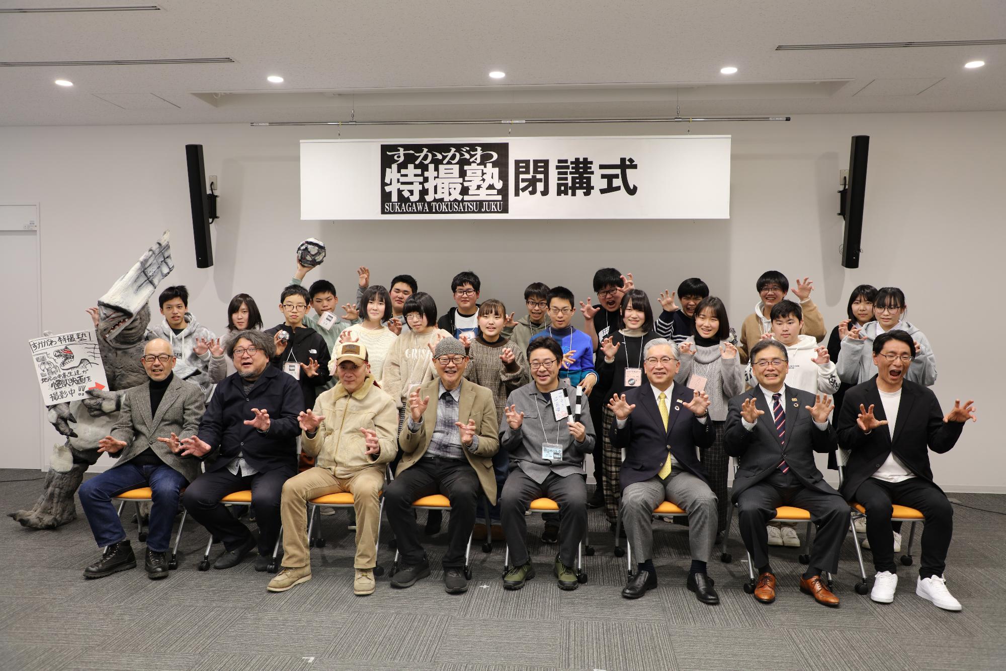 「すかがわ特撮塾閉講式」と書かれた看板の下で、特撮怪獣の着ぐるみや講師、塾生たちが一堂に会し、怪獣の爪を模したポーズで記念撮影をしている写真