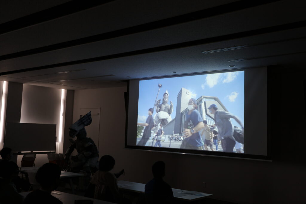 暗い室内で、前方のスクリーンに映し出された特撮映画の上映を鑑賞している写真