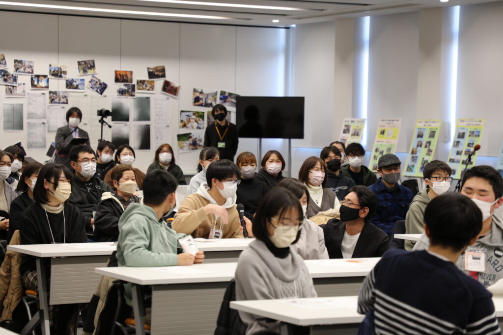 室内で参加者が机に座り、一人の若者が差し出されたマイクに向かって発言する様子を、多くの参加者が耳を傾けている写真