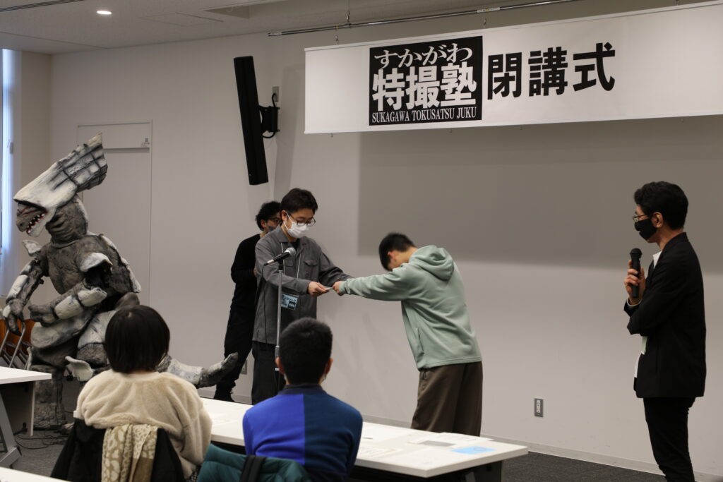 「すかがわ特撮塾 閉講式」の看板が掲げられた会場で、講師から塾生へ卒業証書が手渡され、周囲の参加者たちがその様子を温かく見守っている写真