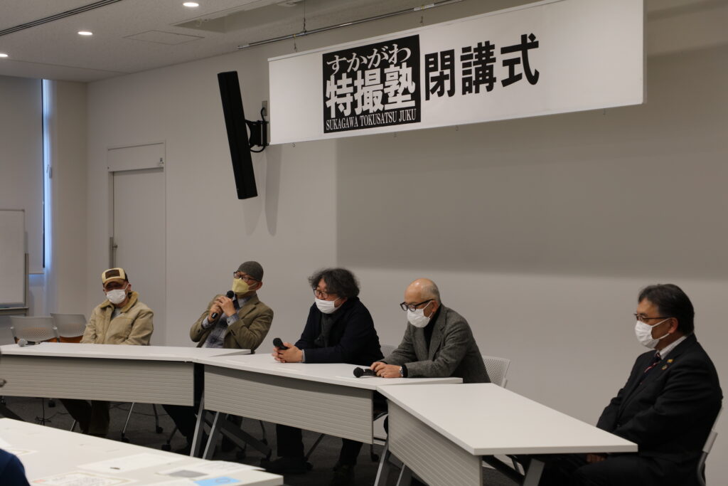 「すかがわ特撮塾 閉講式」と書かれた横断幕が掲げられた会場で、講師陣が白い長机に並んで座り、マイクを手に取りながら話をしている写真