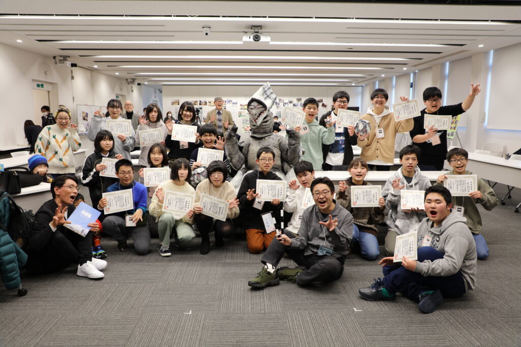 卒業証書を手にした大勢の塾生たちや講師が中央の特撮怪獣を囲むように並び、全員で怪獣ポーズを決めながら写した集合写真