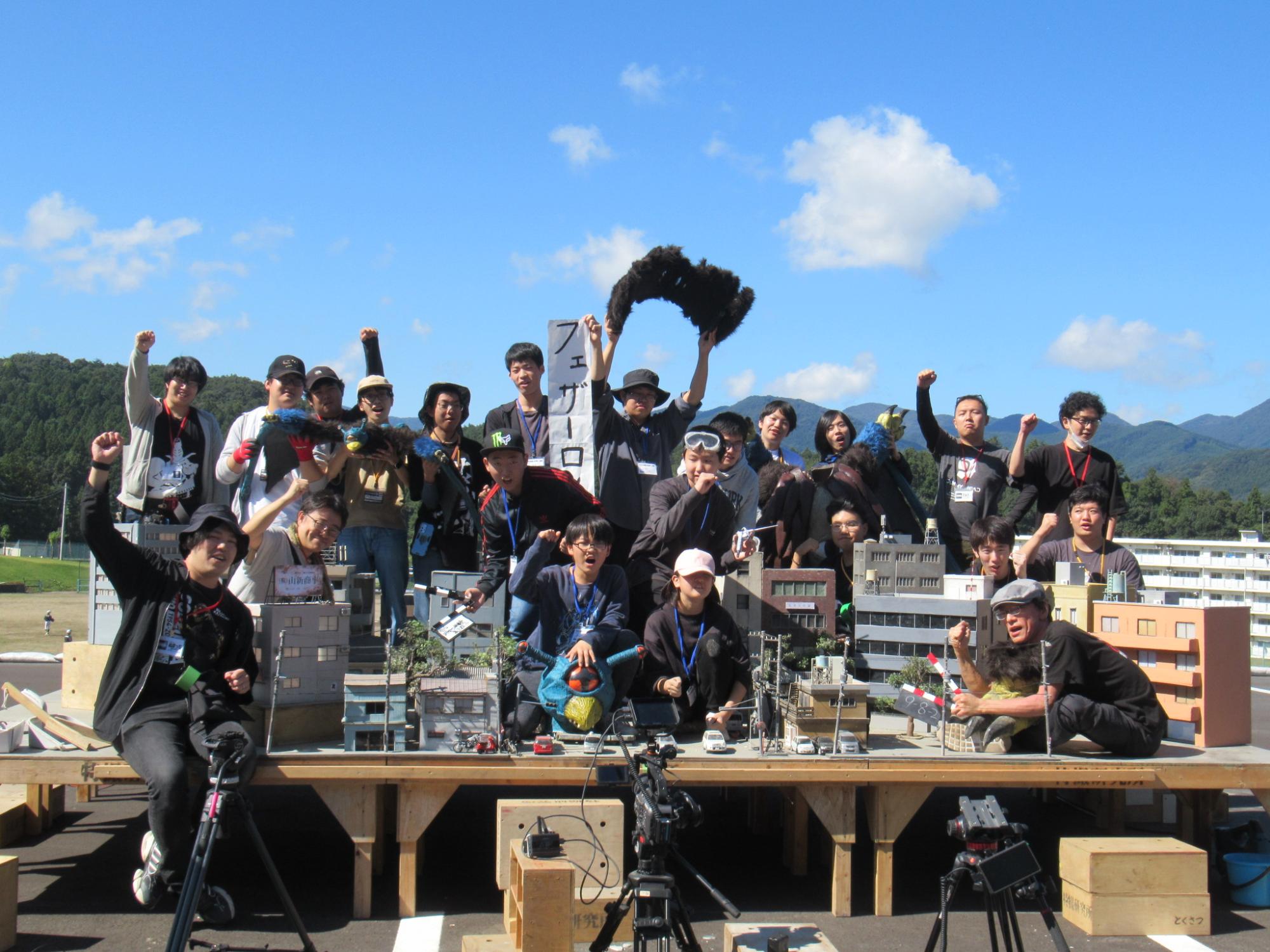 青空の下で多数の参加者がミニチュアの街並みセットを囲み撮影用機材と共にポーズをとる写真