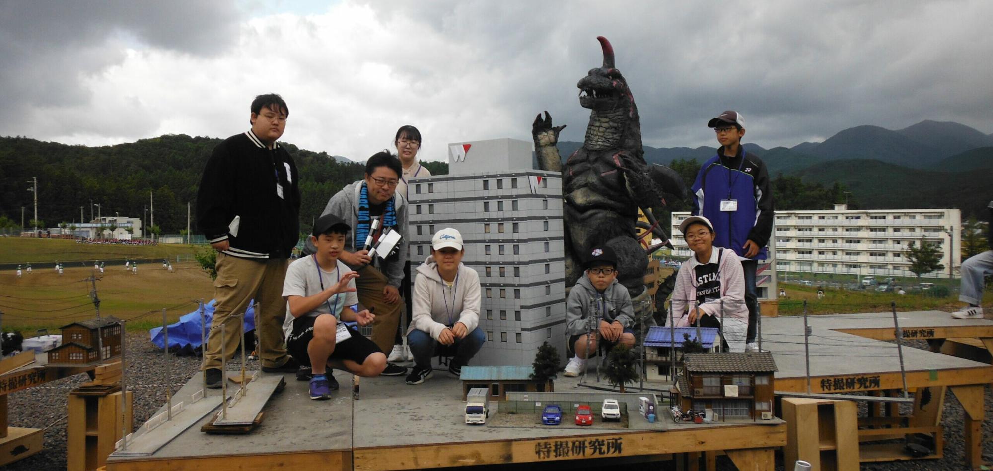 台の上で、大きな怪獣の模型とビル群のジオラマを囲み屋外の山々を背景に講師と塾生が並んでポーズをとっている記念写真