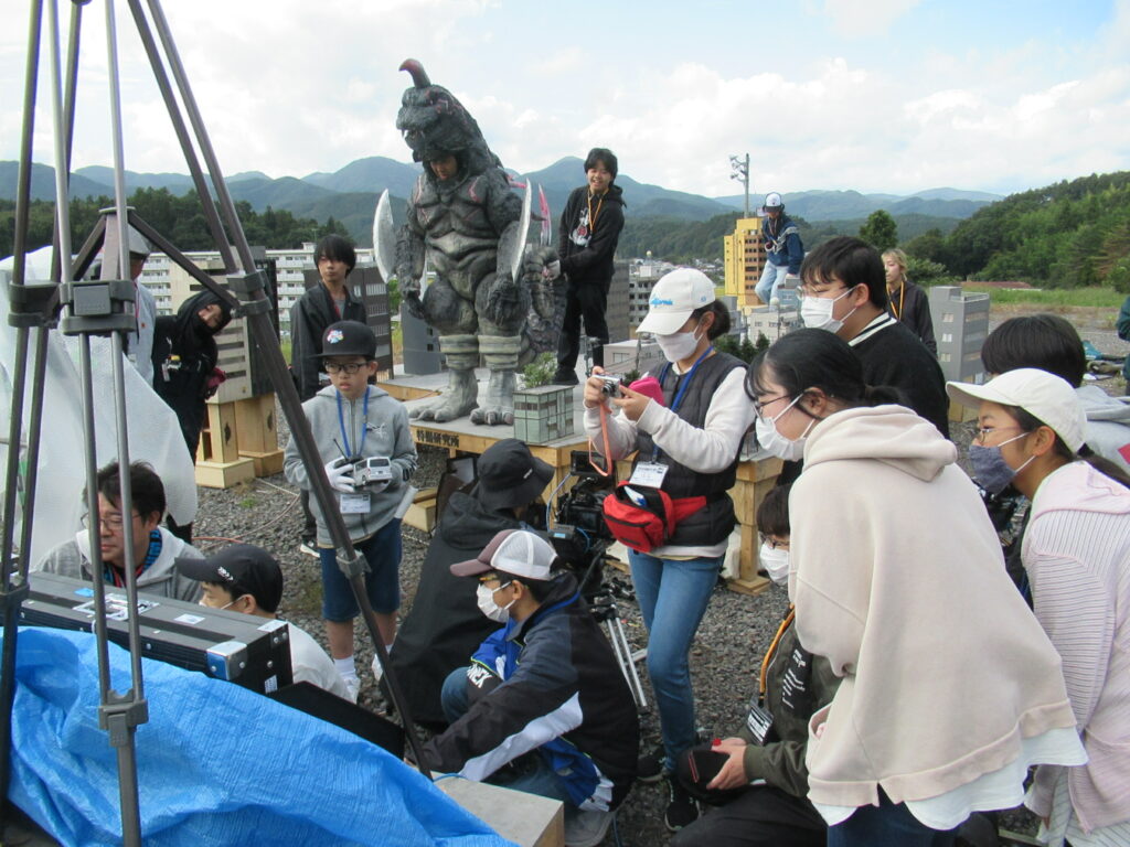 屋外の撮影現場で、ミニチュアの街並みに怪獣が立つセットの前で、大勢のスタッフや塾生たちが撮影した映像や写真を確認している様子の写真