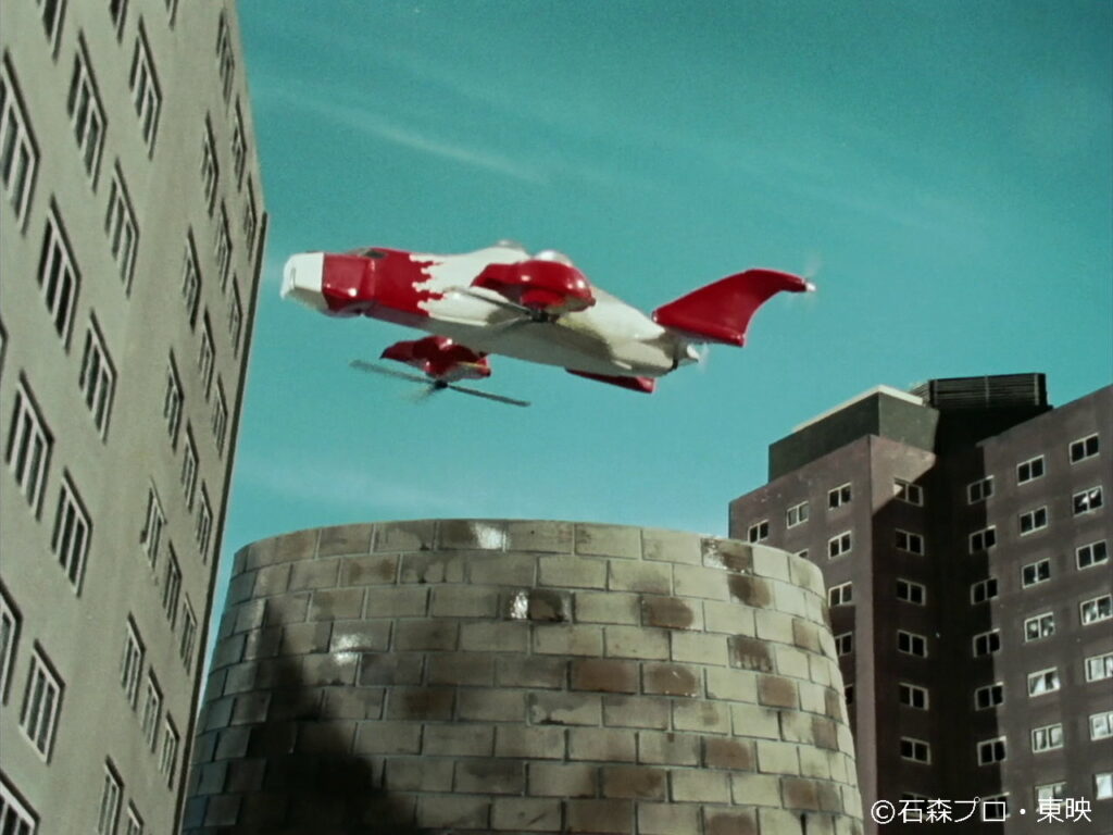 抜けるような青空の下、円筒形の巨大な建造物の影から赤と白のツートンカラーが鮮やかな特殊航空機が飛び立ち、高層ビルが立ち並ぶ都会の街並みの上空を飛行している、特撮作品のワンシーンを切り取った写真