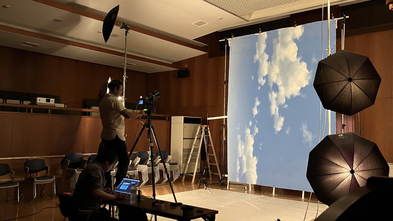 映画撮影のスタジオのような広い室内で、青空と白い雲が大きく描かれた背景パネルを垂直に立て、複数の照明機材や三脚に据えられたカメラ、さらにモニターを確認するスタッフを配置して撮影している写真