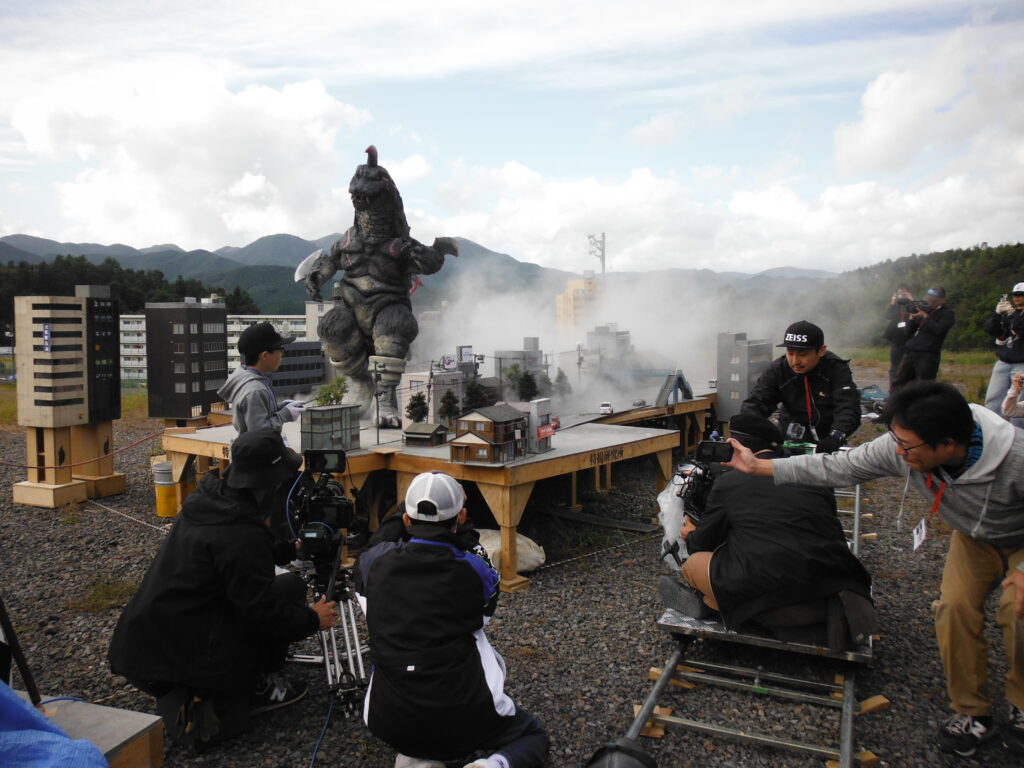 山並みを背景にした屋外の撮影現場で、白煙が立ち込めるミニチュアの街並みに怪獣が立ちが立ち、その手前でカメラマンが撮影し塾生が手伝っている様子の写真