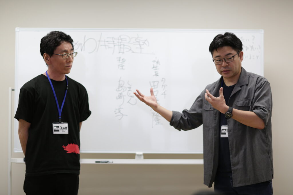 ホワイトボードを背に、黒いTシャツを着た男性が静かに耳を傾ける傍らで、グレーのシャツを羽織った男性が身振り手振りを交えて説明を行っている写真