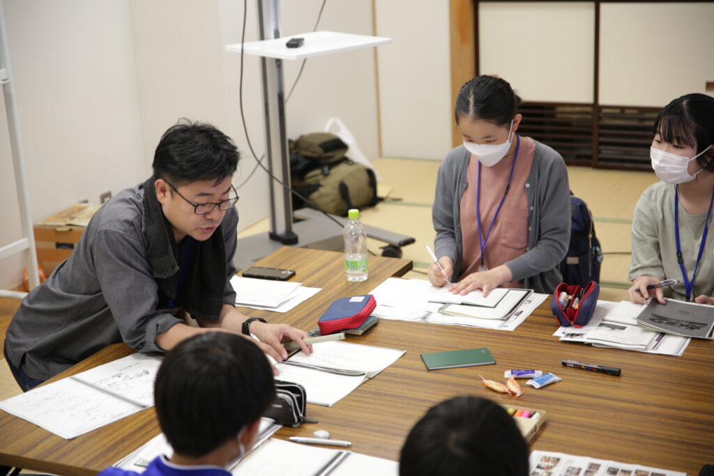 眼鏡をかけた男性講師が木目調のテーブルを囲み、マスクを着用した塾生が熱心にノートを取る写真