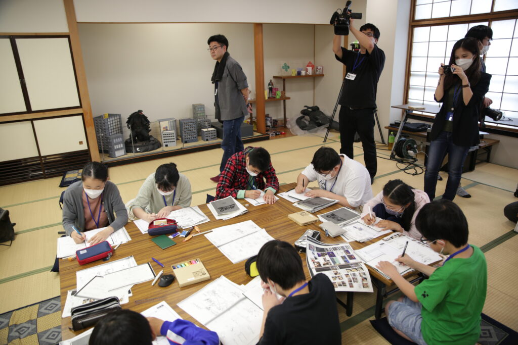 畳敷きの和室で、机を囲んで座る多くの塾生たちが資料を広げて熱心に書き込み作業を行う様子を、ビデオカメラや一眼レフを構えたスタッフが記録している写真