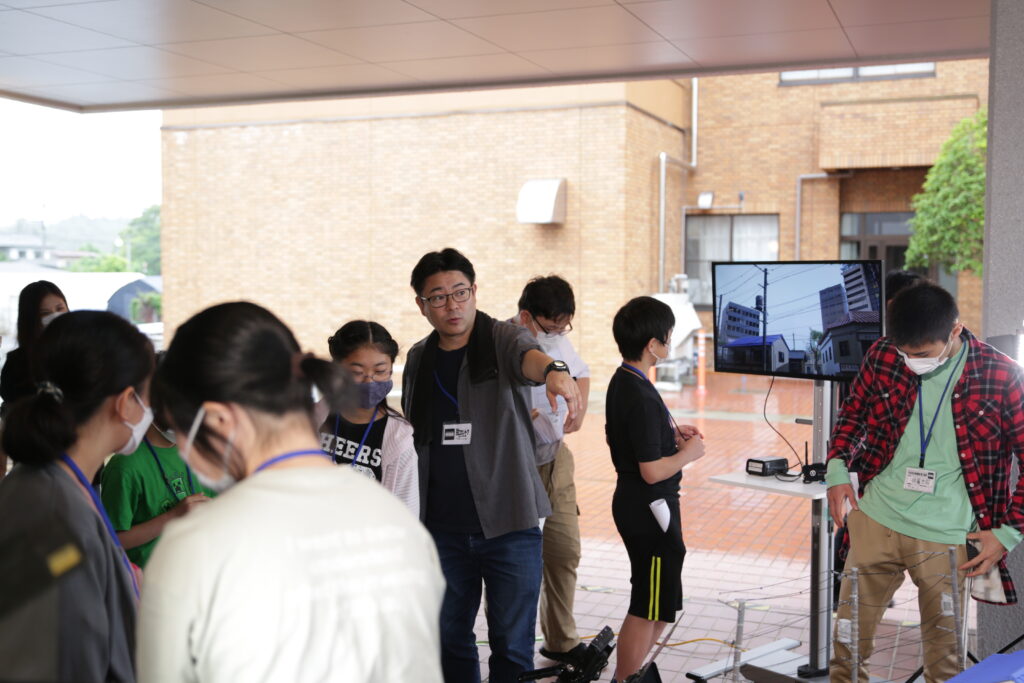 建物の軒下のような屋外空間で、モニターに映し出された映像を確認しながら、男性講師が周囲に集まった若者たちに対して前方を指差し、熱心に解説や指示を行っている写真