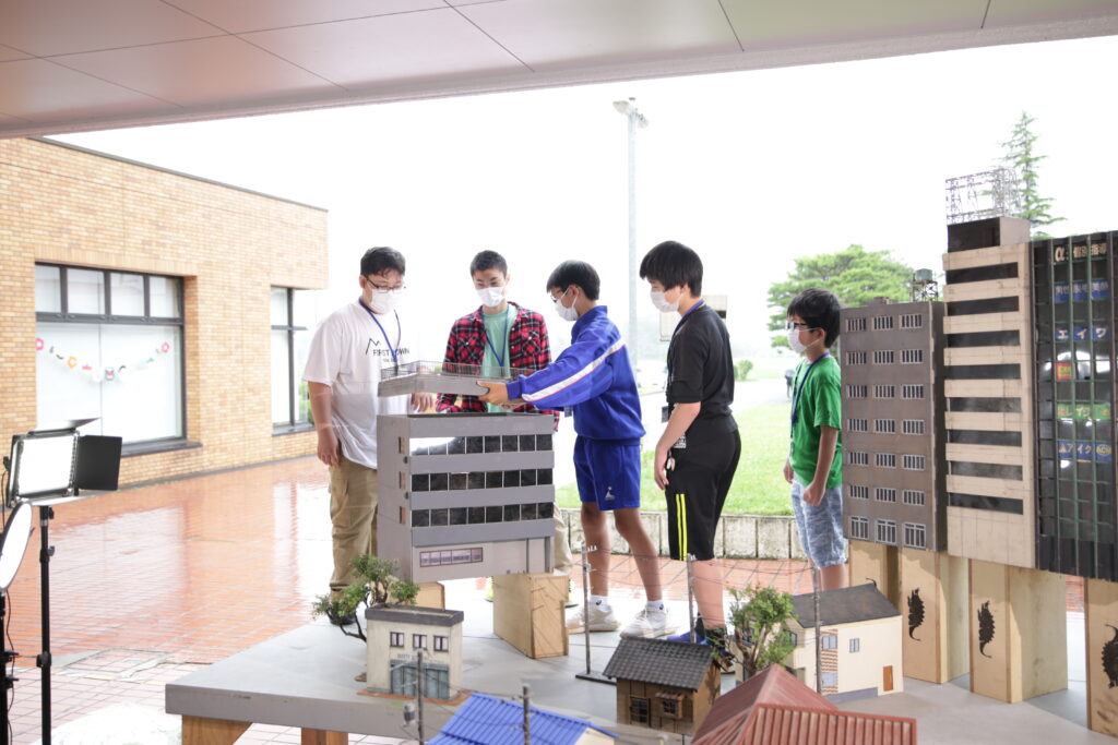 屋外の広場で、マスクを着用した5人の男子塾生たちが、テーブルの上に設置された高層ビルのパーツを慎重に設置している様子の写真