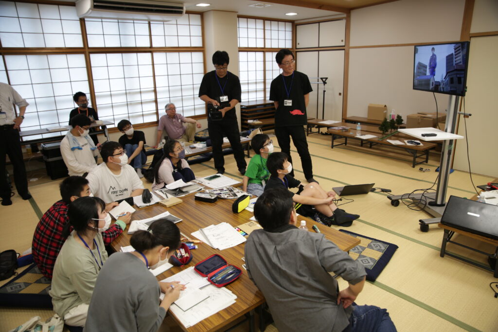 畳敷きの和室で、机を囲んで座る多くの若者たちが資料を広げている中、モニターに映し出された映像を全員で注視している写真