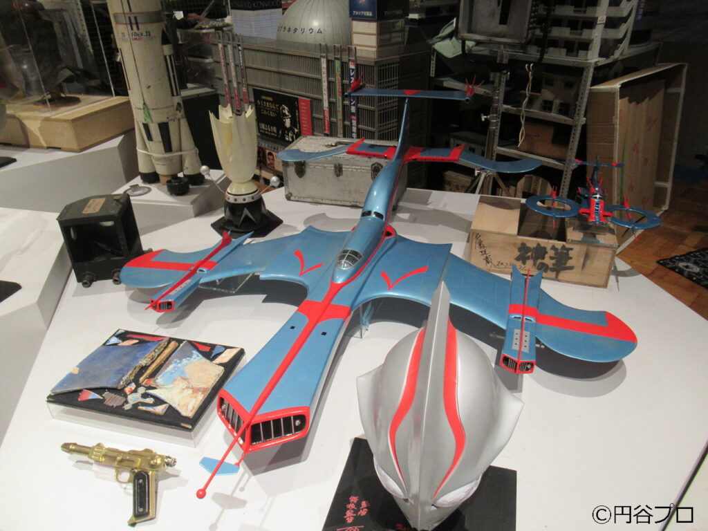 展示台の上に、ウルトラマンのマスクや科学特捜隊のジェットビートル、光線銃などのプロップが整然と並べられ、背後にはビル群やロケットのミニチュアが展示されている写真