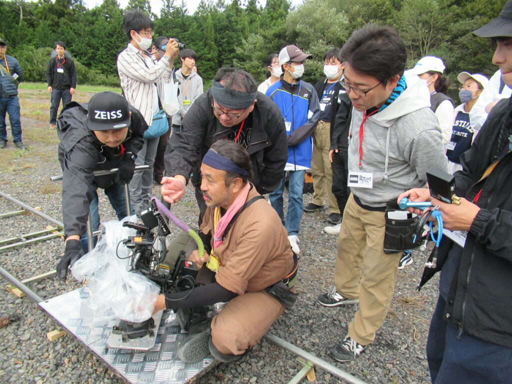 砂利敷きの広場にて、レールの上に設置された撮影用カメラを囲むようにスタッフたちが集まり話をしているのを塾生たちが後ろで見守っている様子の写真