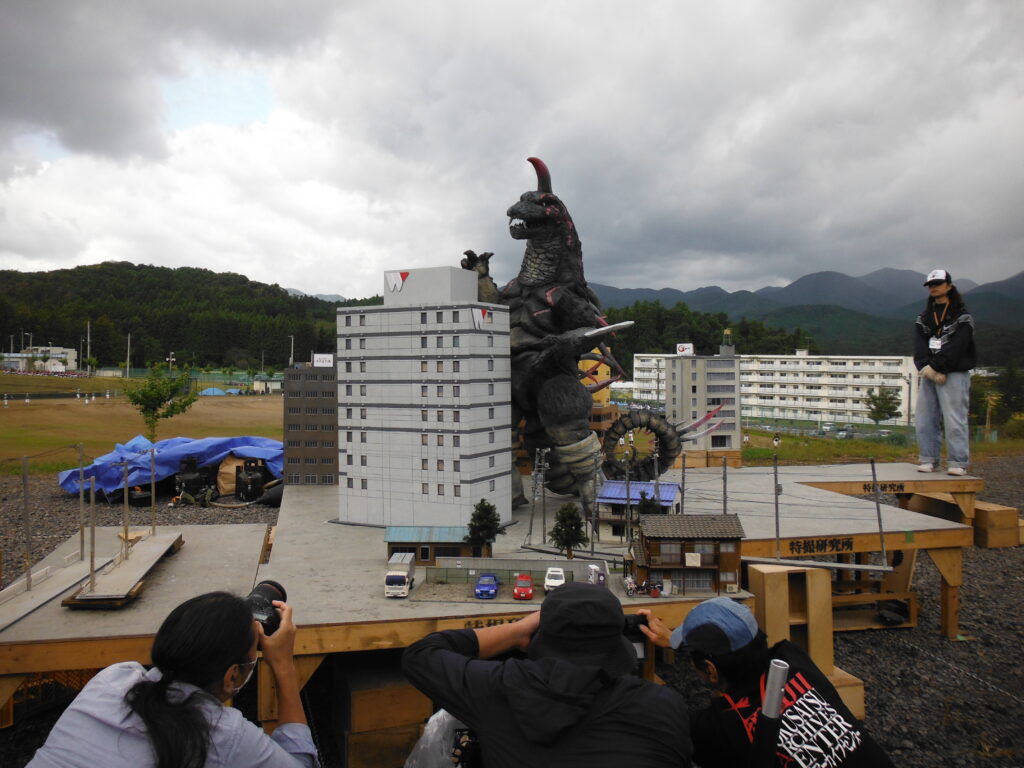 木製の土台に、巨大な怪獣の模型とビルや民家のジオラマが設置され、手前でしゃがみ込んだ塾生やスタッフたちが真剣な表情でレンズを向けて撮影している写真