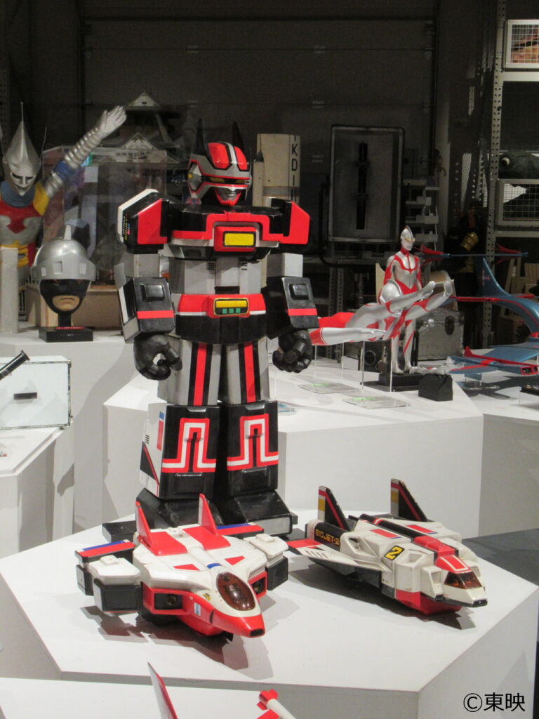 展示台の上に、黒と赤を基調とした巨大ロボットの立像と、その手前に2機の戦闘機が並べられ、周囲には他のヒーローのマスクや模型も見える、特撮作品の造形物を一堂に集めた展示会の写真