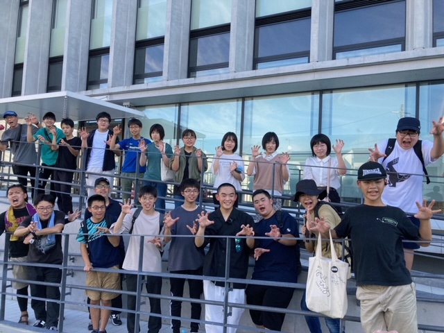 須賀川市役所の建物の外にあるスロープの手すり沿いに、大勢の若者たちが二段に分かれて並び、カメラに向かって一斉に怪獣のようなポーズを笑顔で決めている集合写真