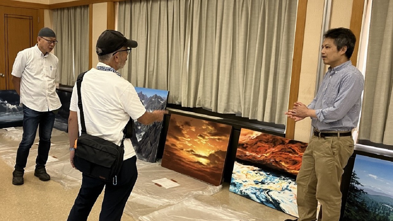 明るい室内で三人の男性が、床に並べられた夕焼け空や荒れ狂う波、険しい山脈などを描いた迫力ある複数の背景画を前にして、身振り手振りを交えながら会話をしている写真