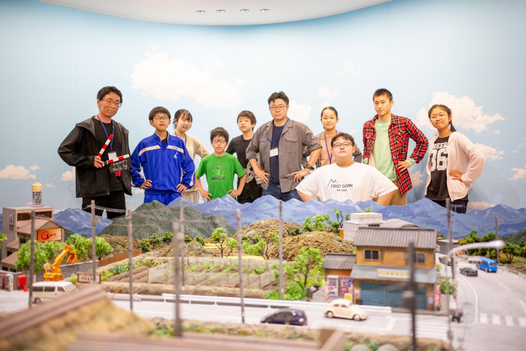 青い空と白い雲の背景を背に、手前の精巧な街並みのミニチュアセットの背景の後ろに総勢10名の男女が立ち並び、カチンコを持つ男性や腰に手を当てたポーズをしている写真