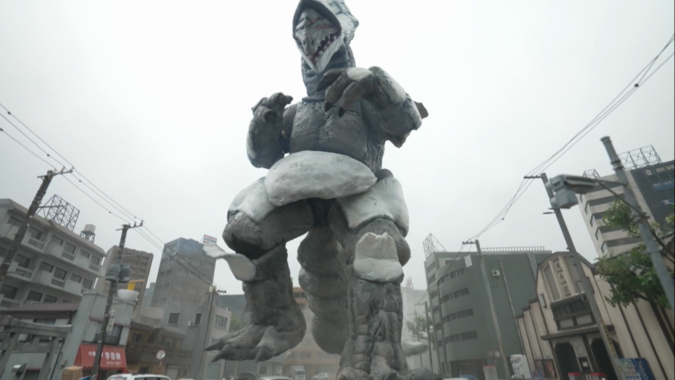 曇り空の下、ビルや電柱が立ち並ぶ市街地の中心に、白とグレーの皮膚を持ち、鋭い牙が見える頭部と強靭な四肢を備えた巨大な二足歩行の怪獣が立ちはだかり、周囲を圧倒するような力強い姿を捉えた実写映像のキャプチャ写真
