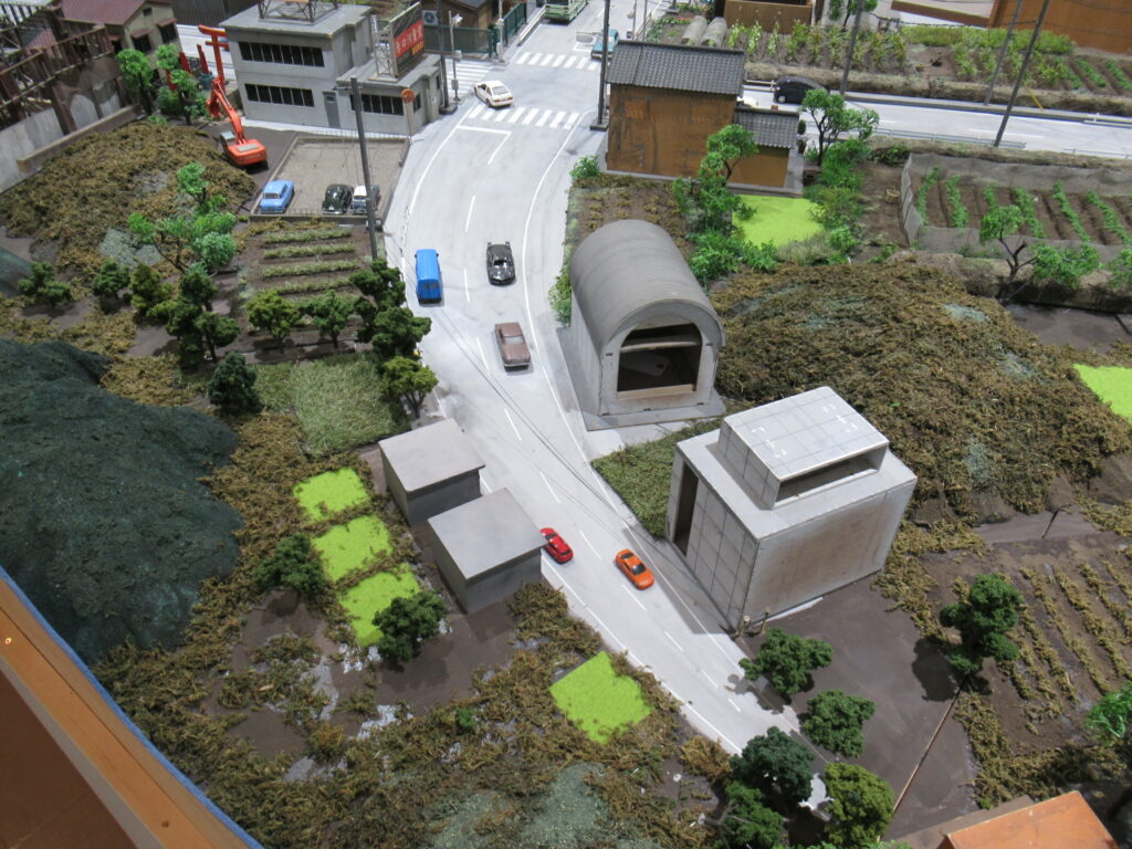 緩やかにカーブする道路沿いに建つ小さな家屋や蔵、畑、そして走行する色とりどりのミニカーが配置されたミニチュアセットの写真