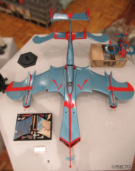 円谷プロの特撮作品に登場する大型戦闘機「スカイホエール」を上空から見下ろした視点で捉えており、青い機体に赤いラインの模様が入った独特な形状の機体模型が白い展示台の上に置かれているミニチュアの写真